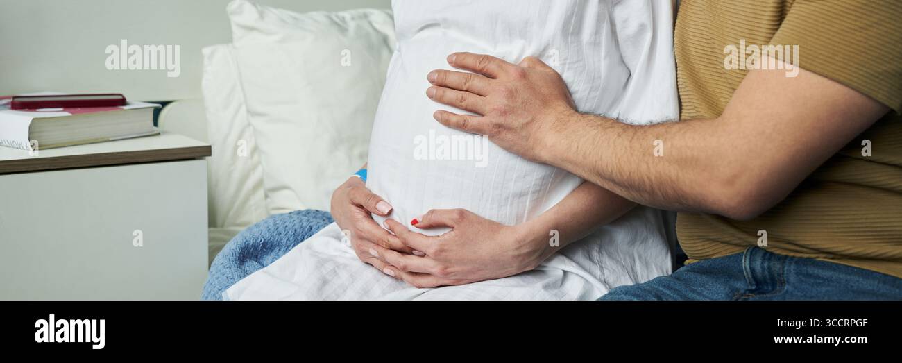 Kaukasische schwangere Frau, die auf dem Bett sitzt und von einem kaukasischen Mann umarmt wird. Beide halten ihren Bauch mit sanft ausgelegten Händen und zeigen einen intimen Moment während der Schwangerschaft zu Hause Stockfoto