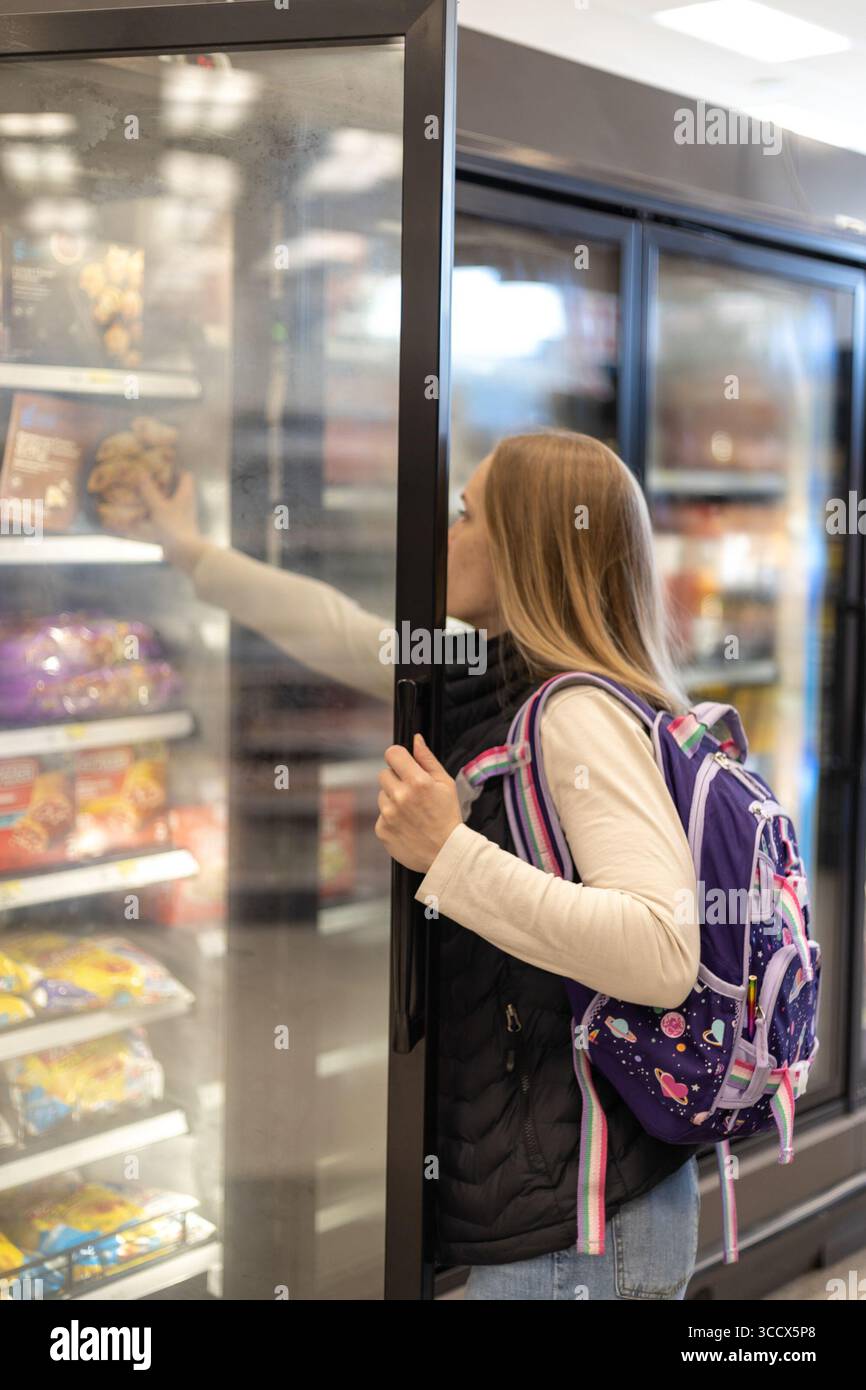 Eine junge Frau mit einem Rucksack öffnet eine Glastür eines kommerziellen Kühl- oder Gefrierschranks in einem Geschäft und greift nach einem Produkt Stockfoto