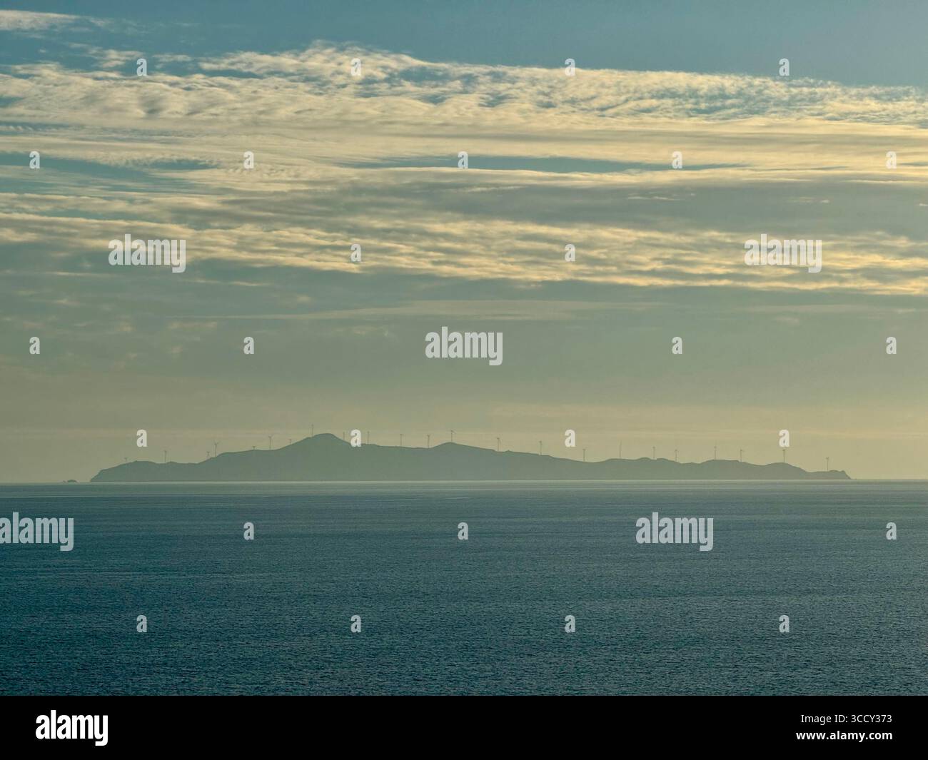 Die unbewohnte Insel Agios Georgios in Griechenland mit ihrem bedeutenden Windpark, der in der Ferne sichtbar ist, umgeben von der Ägäis. - Smartphone-aufgenommenes Stockfoto