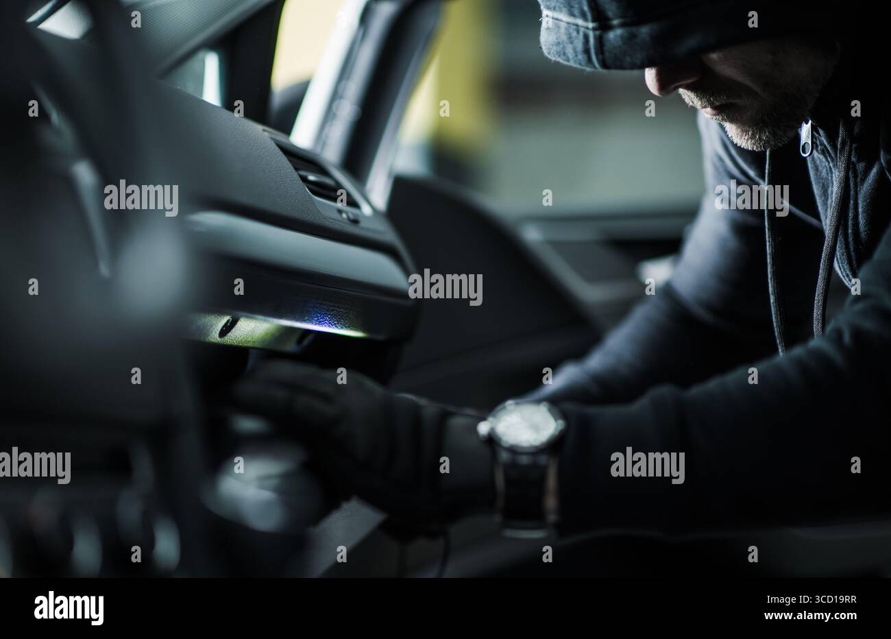 Eine Person mit Motorhaube und Handschuhen sucht bei schlechten Lichtverhältnissen im Armaturenbrett eines Fahrzeugs nach einem potenziellen Diebstahl oder Einbruchversuch. Stockfoto