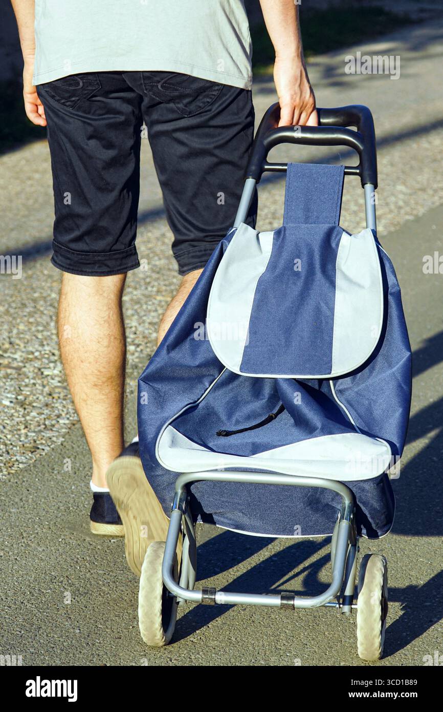 Mann, der mit dem Trolley auf der Straße läuft. Kostengünstiges Einkaufen, Masseneinkäufe auf lokalen Märkten und praktische Lösungen für den Transport schwerer Lebensmittel Stockfoto