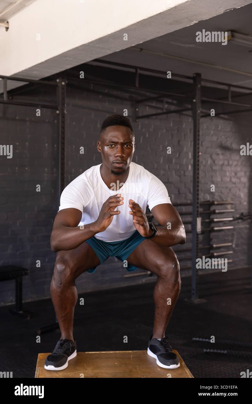 Afroamerikanischer Mann hockt auf plyometrischer Box im Fitnessraum mit Hockgestell, Hantelplatten Stockfoto