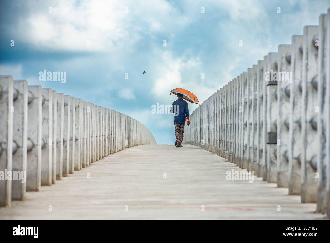 Bogura, Bangladesch - 08. September 2019: Blick auf eine Einzelfigur mit orangefarbenem Regenschirm, die eine Fußgängerbrücke aus Beton unter bewölktem Himmel hinaufsteigt. Stockfoto