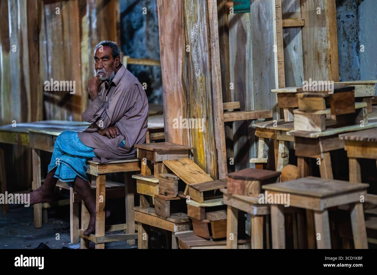 Bogura, Bangladesch – 21. März 2022: Blick auf einen nachdenklichen Handwerker inmitten eines Meeres aus Holzstühlen und -Tischen, sein verwittertes Gesicht zeugt von der jahrelangen Holzformung in seiner Werkstatt. Stockfoto