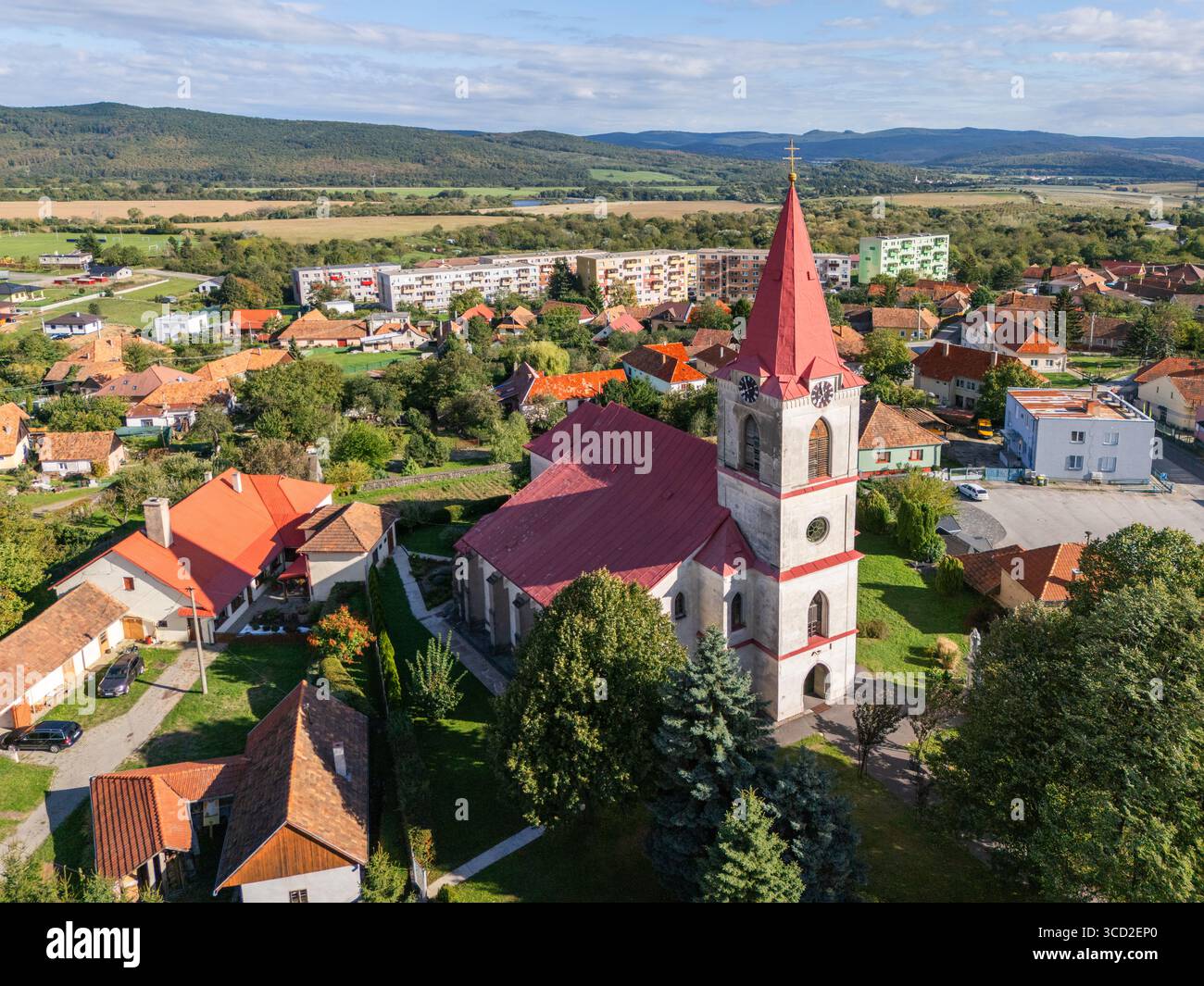 Aus der Vogelperspektive einer Kirche mit einem markanten roten Dach und einem Turm, der über dem umliegenden Dorf ragt, eingerahmt von fernen Hügeln, Kamenica nad Hronom, SL Stockfoto