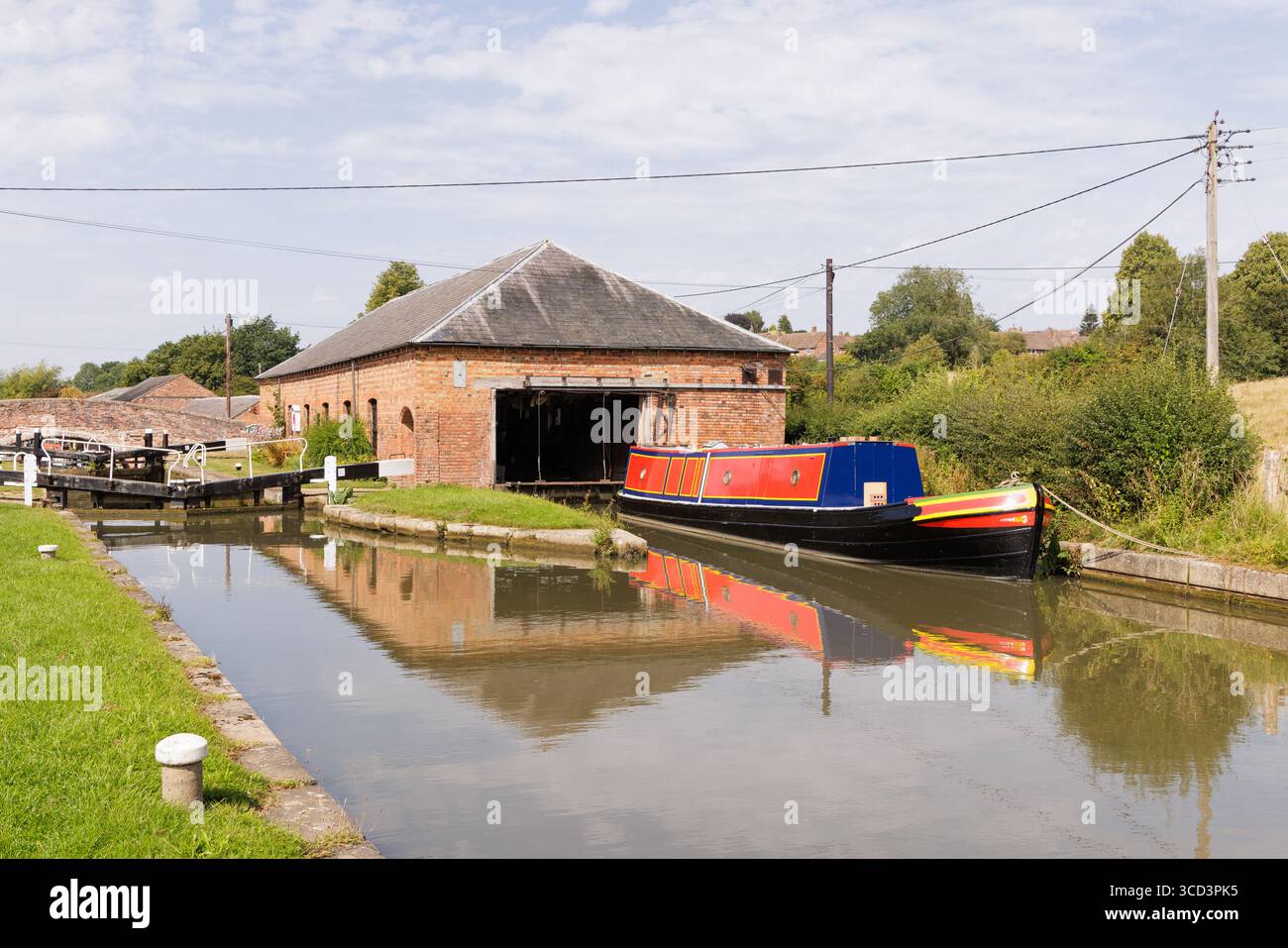 Braunston, Northamptonshire, Vereinigtes Königreich: Ein farbenfrohes Schmalboot, das vor einem alten Werkstattgebäude vor Anker liegt, neben einer Schleuse am Grand Union Canal. Stockfoto