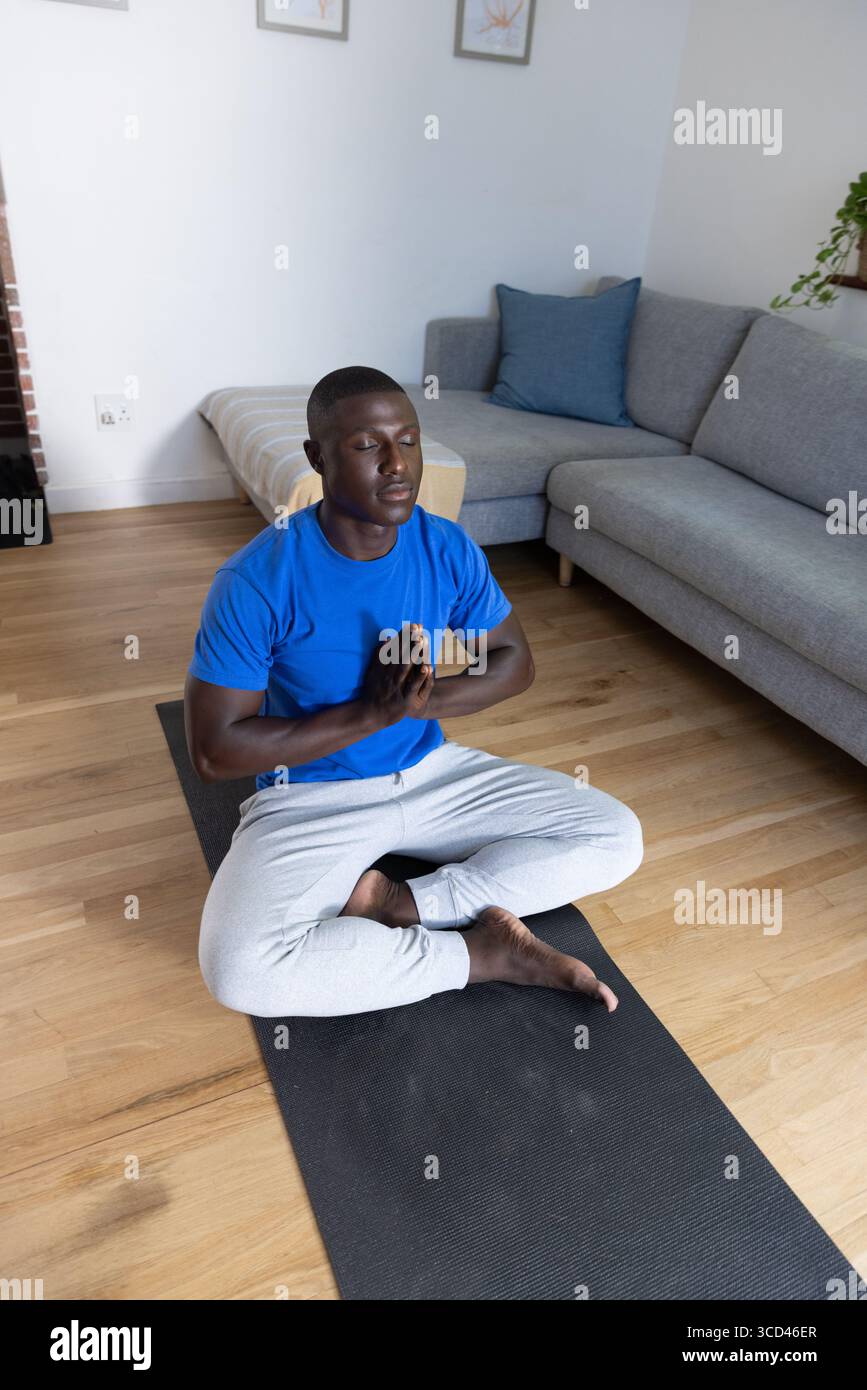 Afroamerikanischer Mann mit Sportbekleidung, der zu Hause auf schwarzer Yogamatte mit botanischen Drucken meditiert Stockfoto