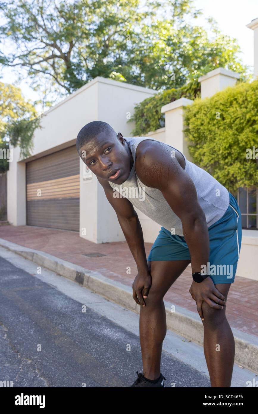 Afroamerikaner, der sich nach vorne beugt und auf der Vorstadtstraße mit Smartwatch und Schuhen Atem schnappt Stockfoto