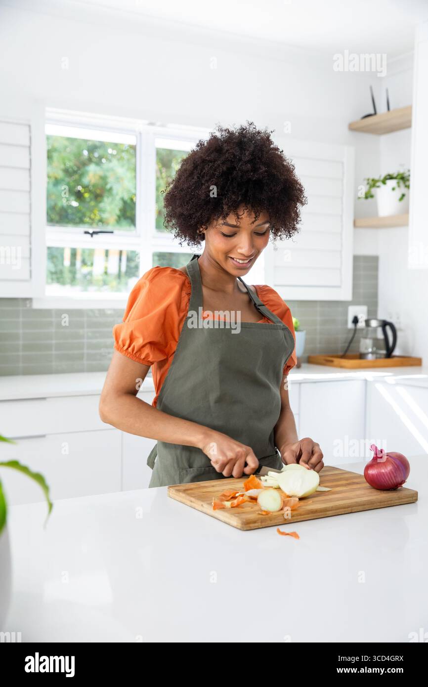 Frau mit orangefarbenem Oberteil und grüner Schürze, die Zwiebel mit Messer an Bord in moderner Küche schneidet Stockfoto