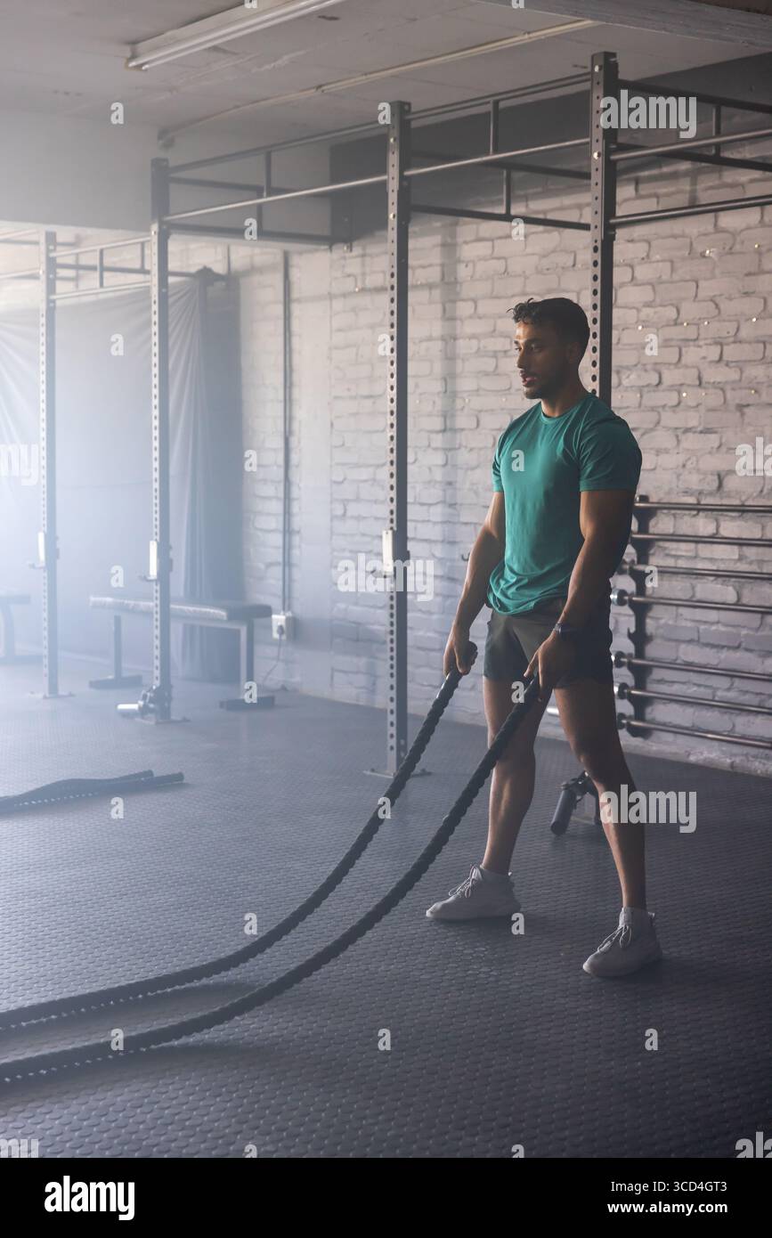 Muskulöser Mann schwingt Kampfseile im Fitnessstudio neben dem Rig, Langhantelständer und Bänken auf Gummimatten Stockfoto