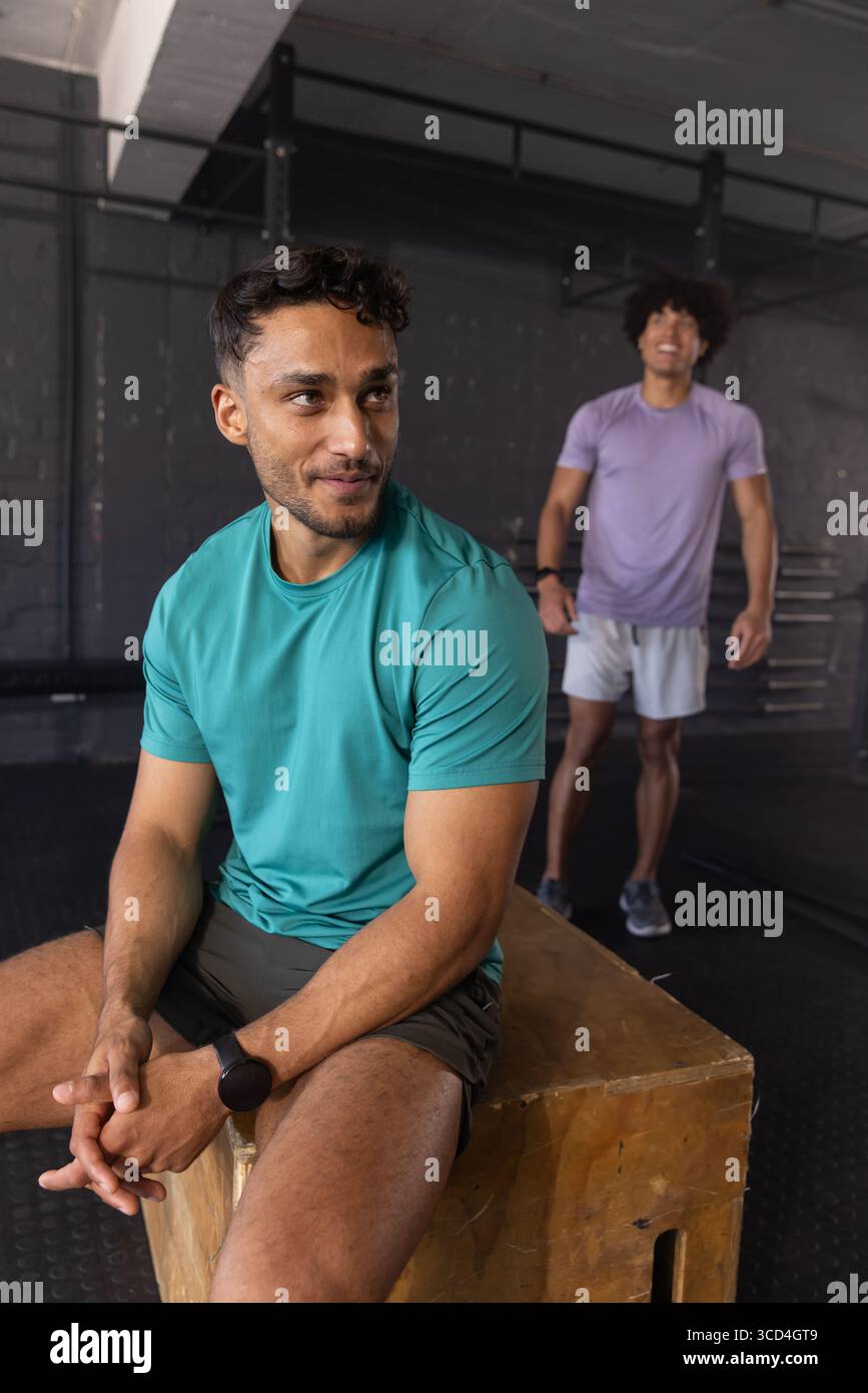 Verschiedene männliche Freunde, die auf einer hölzernen Plyometriekasten im Fitnessstudio neben dem Hockgestell liegen, Kopierraum Stockfoto