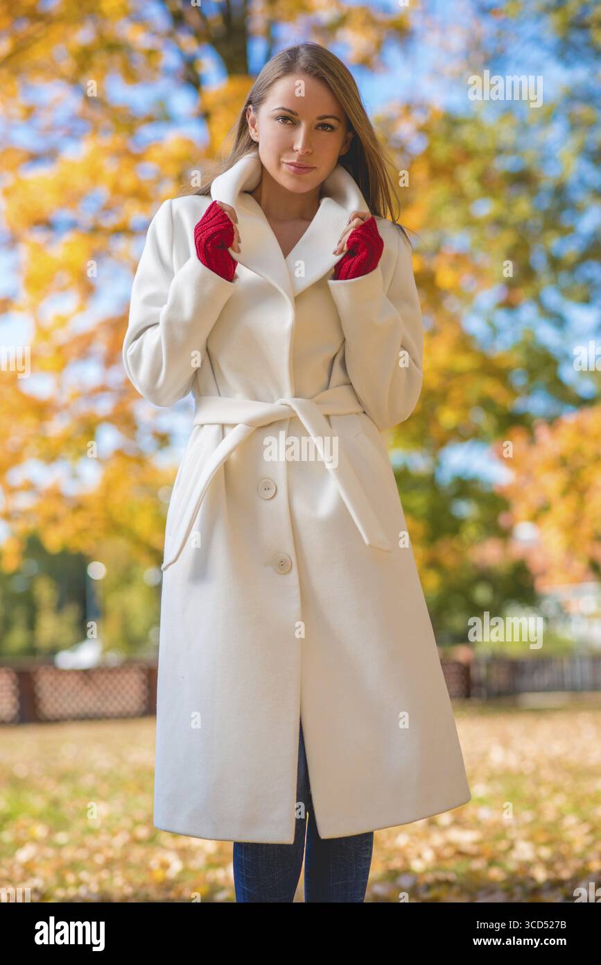 Modische, selbstbewusste junge Frau mit einem stilvollen weißen Mantel und roten Fäustlingen, die draußen in einem farbenfrohen Herbstpark steht und auf die Kamera blickt Stockfoto