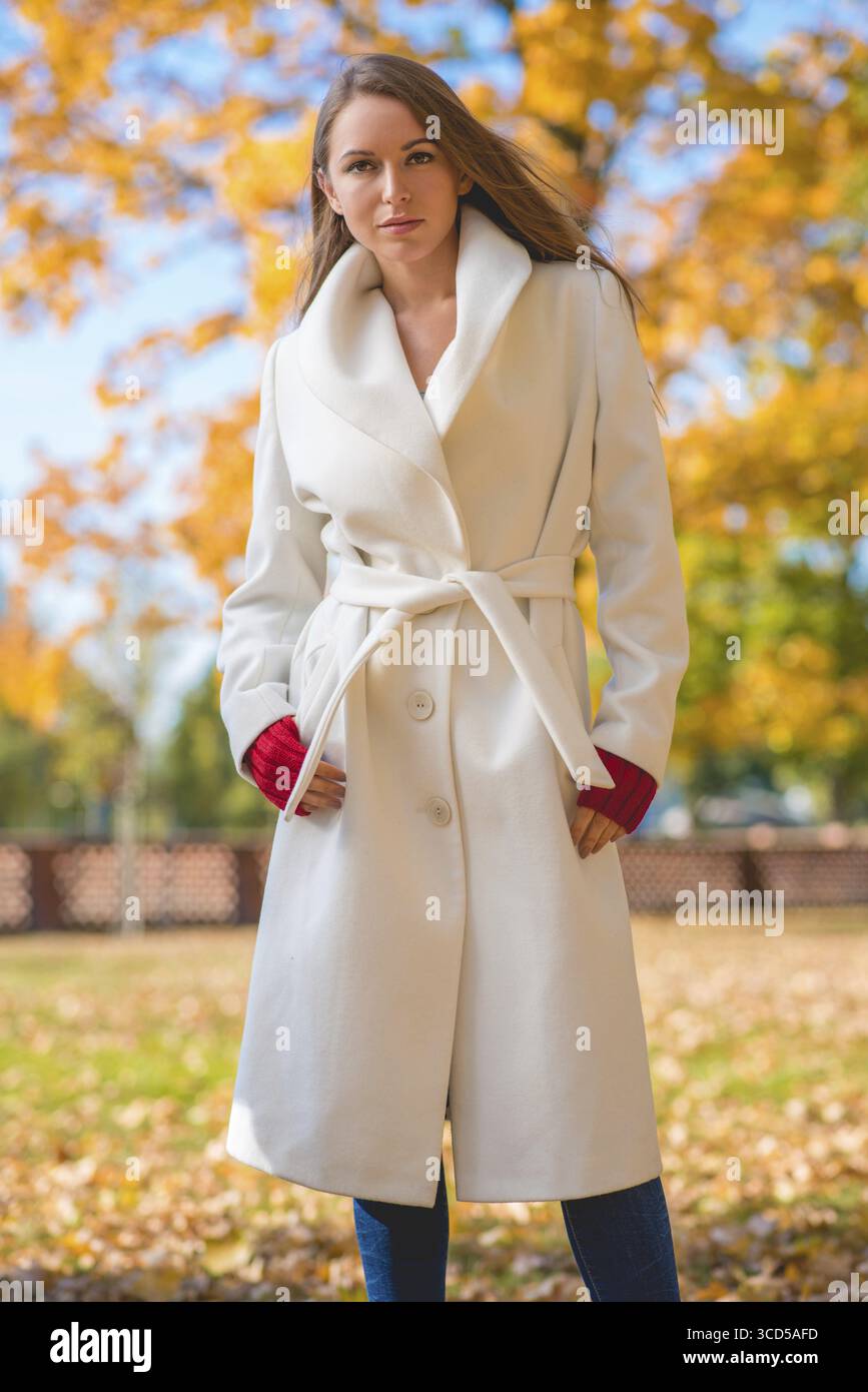 Modische, selbstbewusste junge Frau mit einem stilvollen weißen Mantel und roten Fäustlingen, die draußen in einem farbenfrohen Herbstpark steht und auf die Kamera blickt Stockfoto