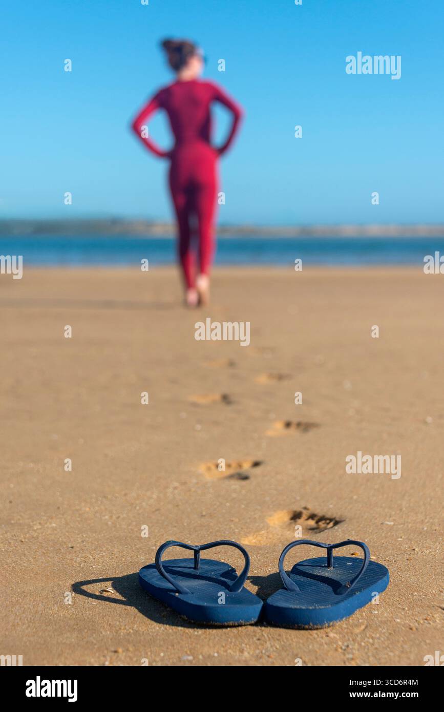 Flip-Flops am Sandy Beach mit einer Frau in Rot, die zum Meer geht Stockfoto