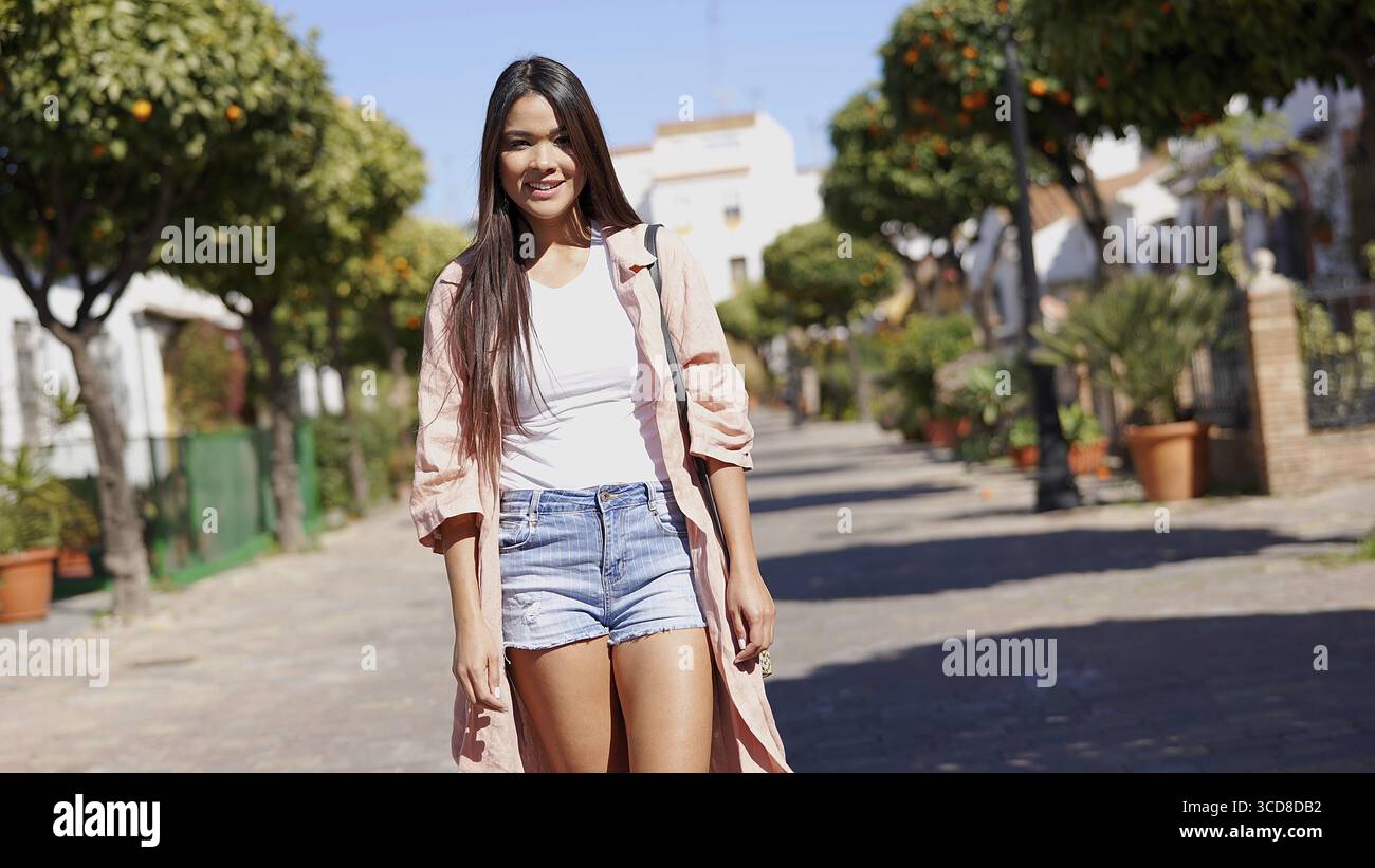 Eine fröhliche junge Frau, die einen gemütlichen Spaziergang entlang einer malerischen Straße in einer sonnigen spanischen Stadt genießt, umgeben von Grün und Orangenbäumen Stockfoto