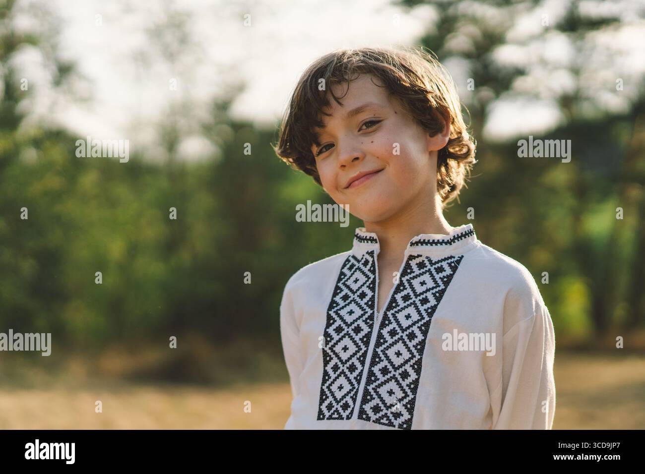 Ein Junge lächelt selbstbewusst, während er auf einem sonnendurchfluteten Feld steht und ein traditionelles besticktes Hemd trägt Stockfoto