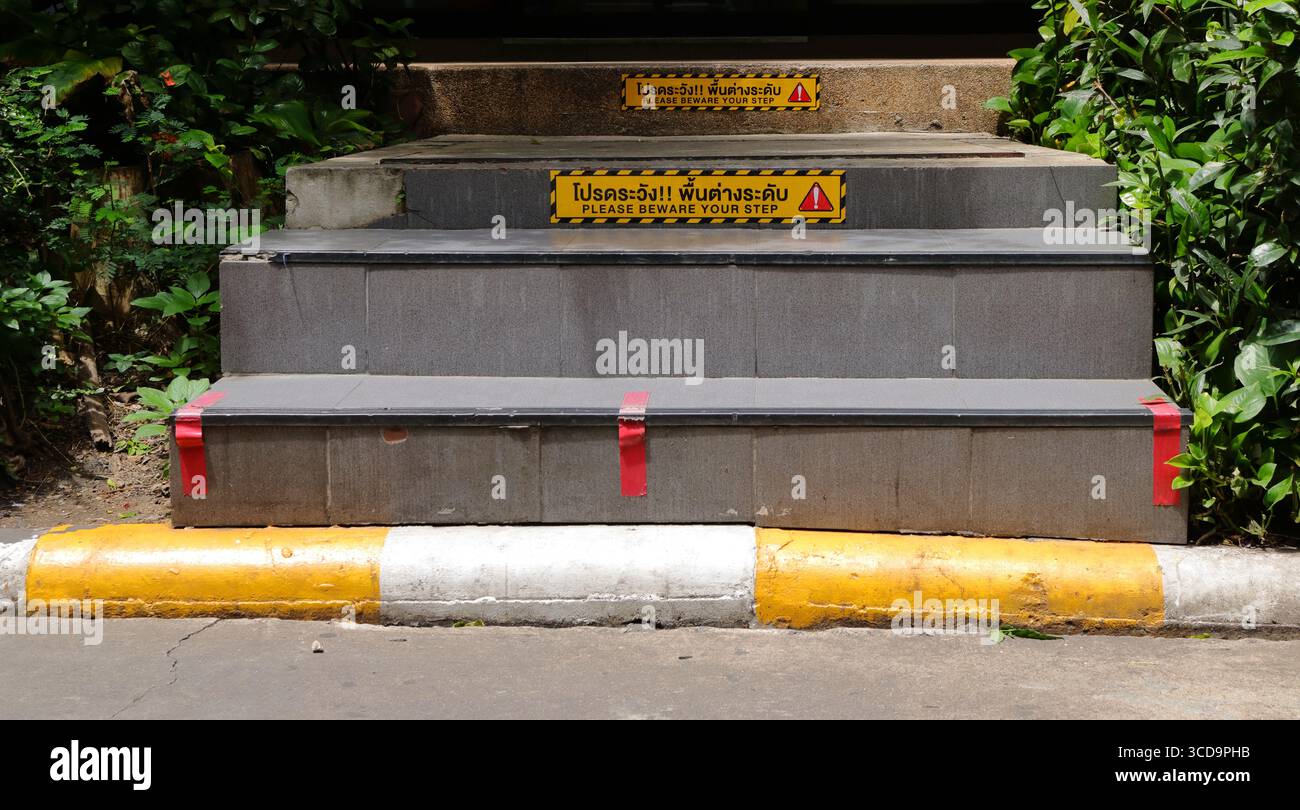 Bitte hüten Sie sich vor Ihrem Treppenschild auf Englisch und Thai, das an der Treppenstufe angebracht ist Stockfoto