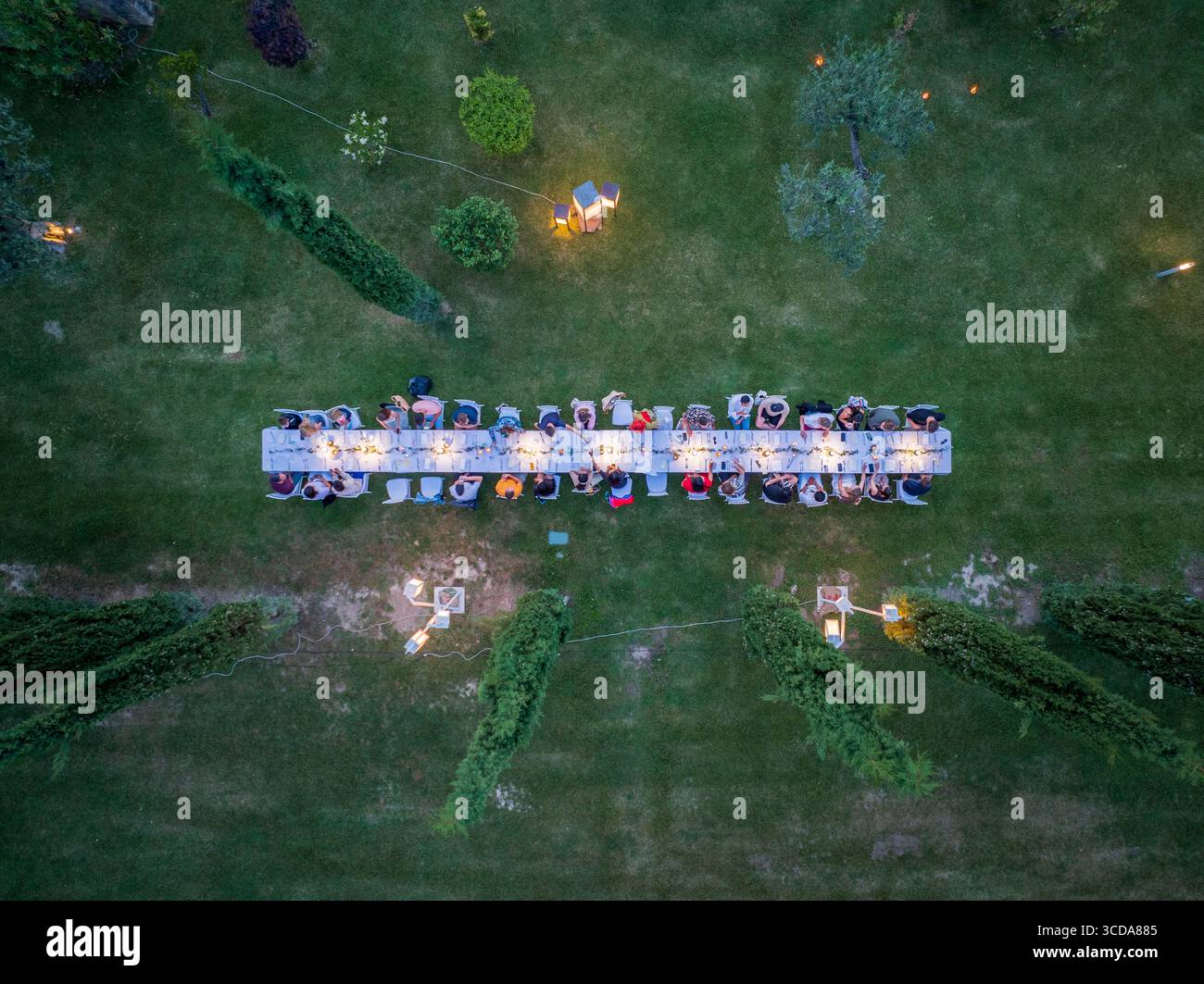 Blick aus der Vogelperspektive auf einen langen Tisch für ein Festmahl, eingebettet zwischen üppig grünen Rasenflächen und Bäumen, beleuchtet von sanften, warmen Lichtern, Assisi, Umbrien, Italien. Stockfoto