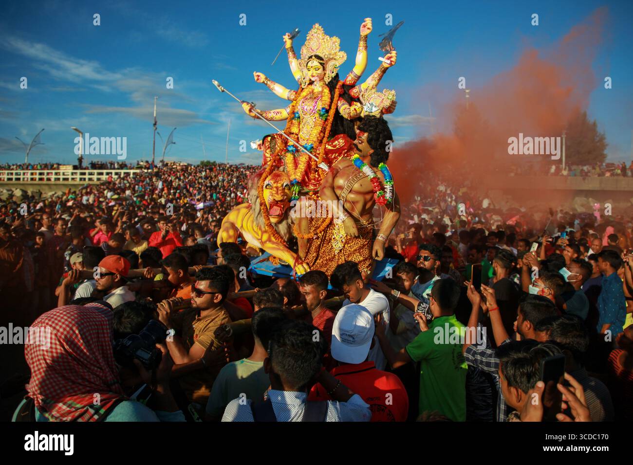 Chittagong, Bangladesch - 5. Oktober 2022: Blick auf ein lebendiges Durga-Idol, das durch eine dichte Menschenmenge getragen wird, die Luft ist von rötlich-orangenem Rauch vor einem hellblauen Himmel bedeckt. Stockfoto