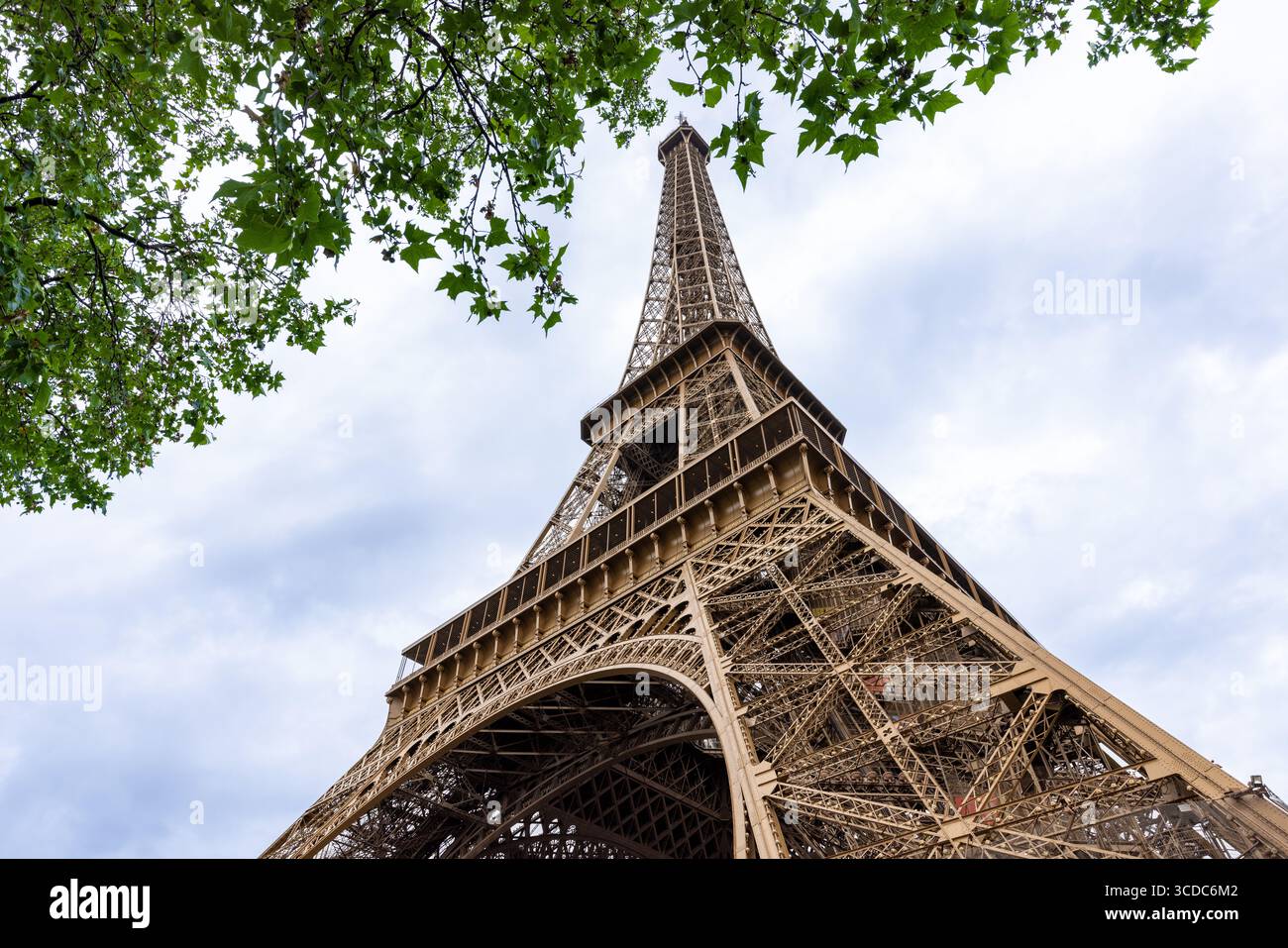 Blick auf das Gittereisen des Eiffelturms, das durch den Himmel ragt, eingerahmt von leuchtenden grünen Blättern vor einer weichen, wolkenbedeckten Kulisse, Paris, Île-de-France, Frankreich. Stockfoto