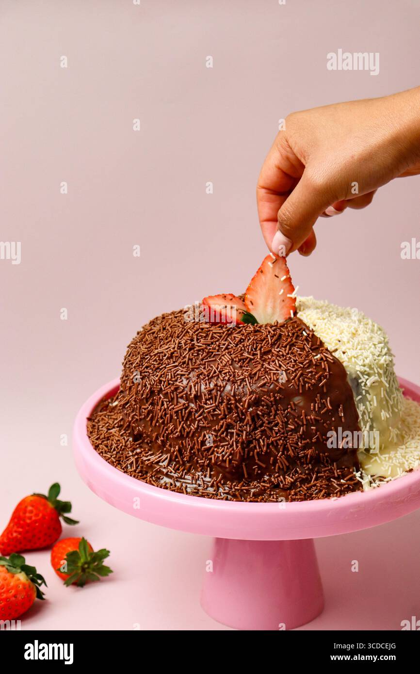 Eine Hand legt eine frische Erdbeerscheibe auf einen Brigadeiro-Kuchen, ein traditionelles brasilianisches Dessert, das halb mit Schokoladenstreuseln bedeckt ist Stockfoto