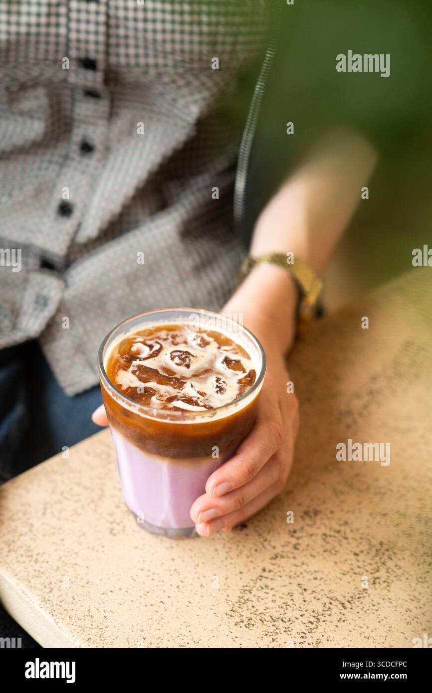 Nahaufnahme einer Frauenhand, die ein Glas Lavendelkaffee in einem modernen Café hält. Kaltgebräu mit blumigem Aroma, sanften Pastelltönen. Stockfoto
