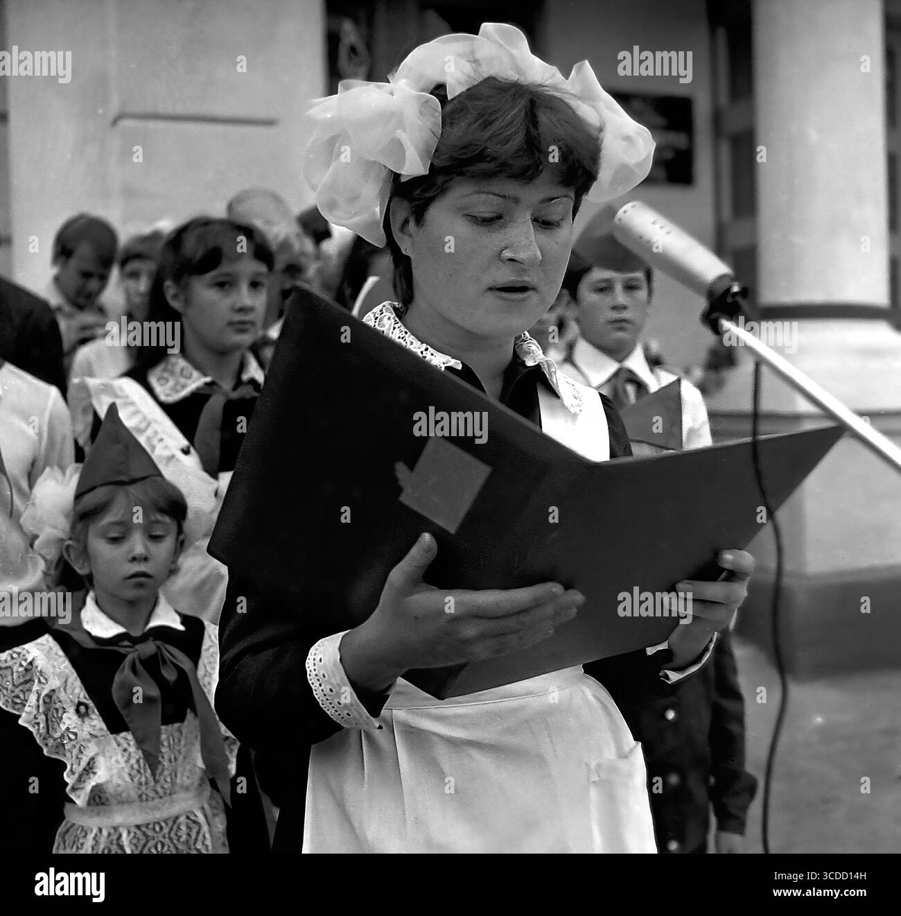 Ein Schulmädchen in formaler sowjetischer Uniform mit Bögen liest während der feierlichen Versammlung zum Tag des Wissens in der Schule Nr. 8 in Sloviansk, 1984 Gedichte oder einen Gruß am Mikrofon. Stockfoto