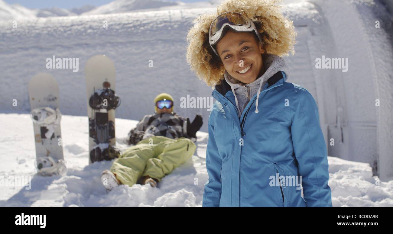 Niedlicher Skifahrer mit blondem Afro und blauer Jacke, der neben dem Freund steht und Snowboards im Schnee stecken Stockfoto