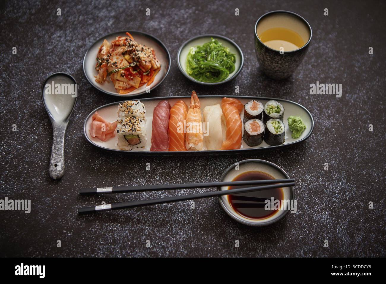 Asiatische Küche. Verschiedene Sushi-Rollen auf Keramikplatten. Kimchi und goma Wakame Salate. Sojasuppe und Essstäbchen an den Seiten. Grungy Dark bac Stockfoto