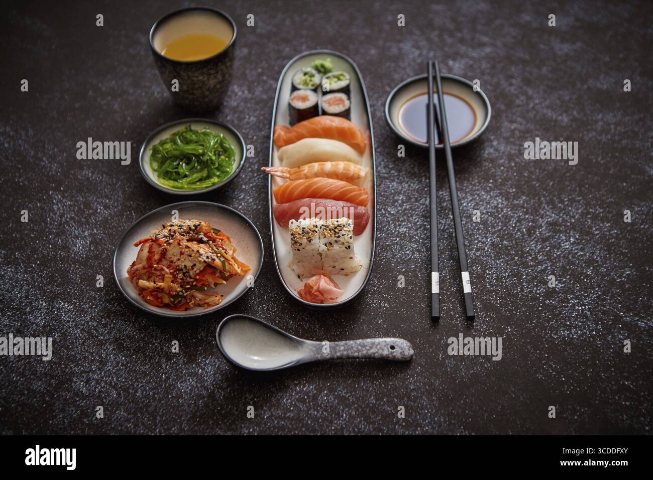 Asiatische Küche. Verschiedene Sushi-Rollen auf Keramikplatten. Kimchi und goma Wakame Salate. Sojasuppe und Essstäbchen an den Seiten. Grungy Dark bac Stockfoto
