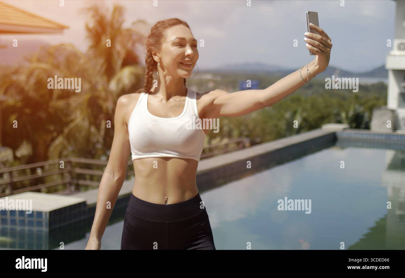 Glückliche schlanke Frau in Sportkleidung lächelt und macht Selfie gegen den Swimmingpool während des Fitnesstrainings an sonnigen Tagen im Resort Stockfoto
