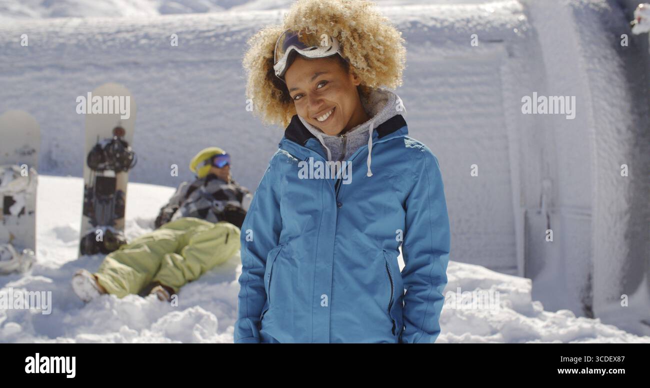 Niedlicher Skifahrer mit blondem Afro und blauer Jacke, der neben dem Freund steht und Snowboards im Schnee stecken Stockfoto