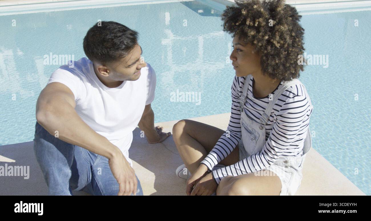 Young African American Paar mit einem schweren Chat sitzen zusammen in der heißen Sommersonne auf der gepflasterten Umrahmung eines Schwimmbades Stockfoto