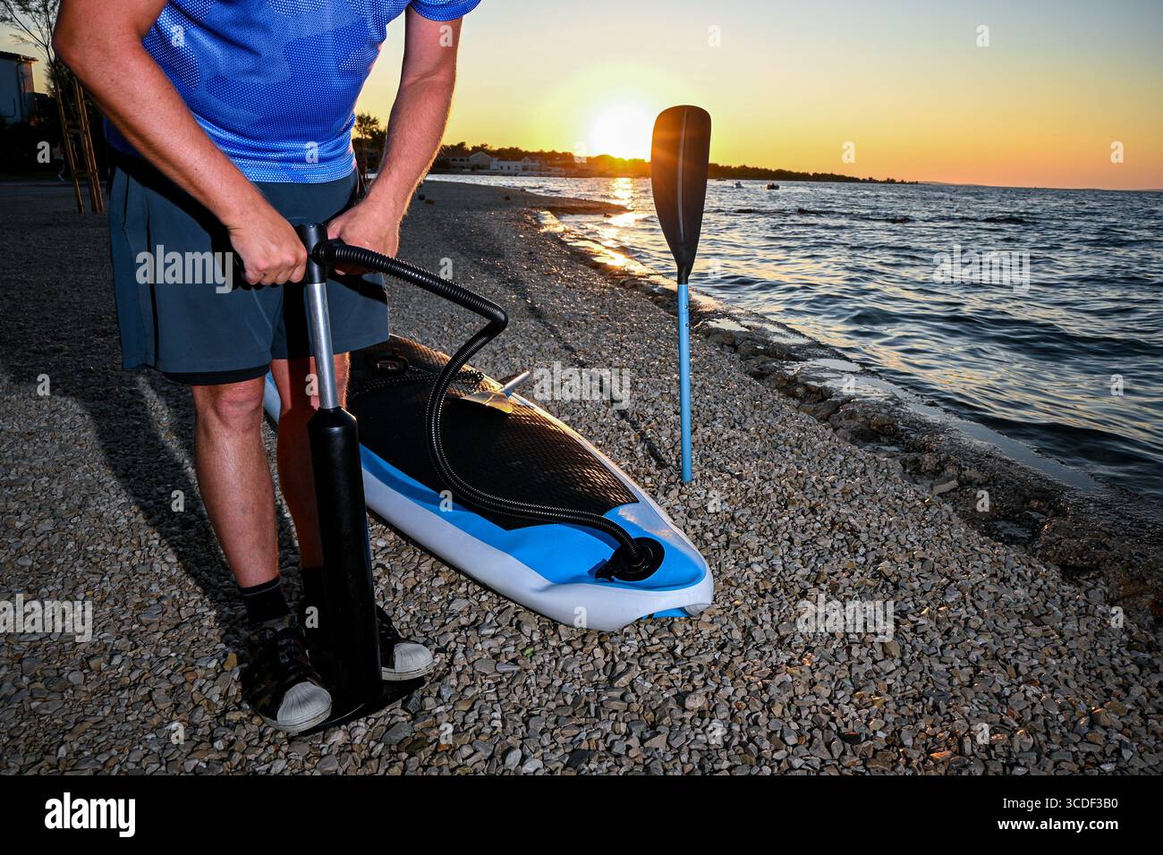 Sportler, der sein Sup Board mit einer Handpumpe an einem Kieselstrand bei Sonnenuntergang aufbläst und sich auf ein entspannendes Paddeln auf dem ruhigen Wasser vorbereitet. Stockfoto