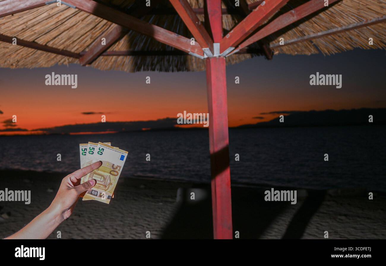 Hand hält Euro-Banknoten an einem Strand mit wunderschönem Sonnenuntergang im Hintergrund. Stockfoto