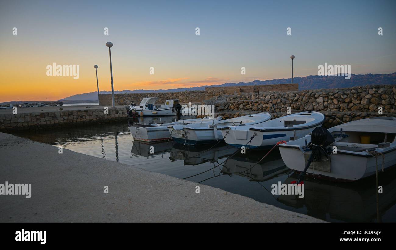 Kleine weiße Boote, die im ruhigen Hafen verankert sind und sich bei Sonnenuntergang auf ruhigem Wasser mit Bergketten im Hintergrund spiegeln. Stockfoto