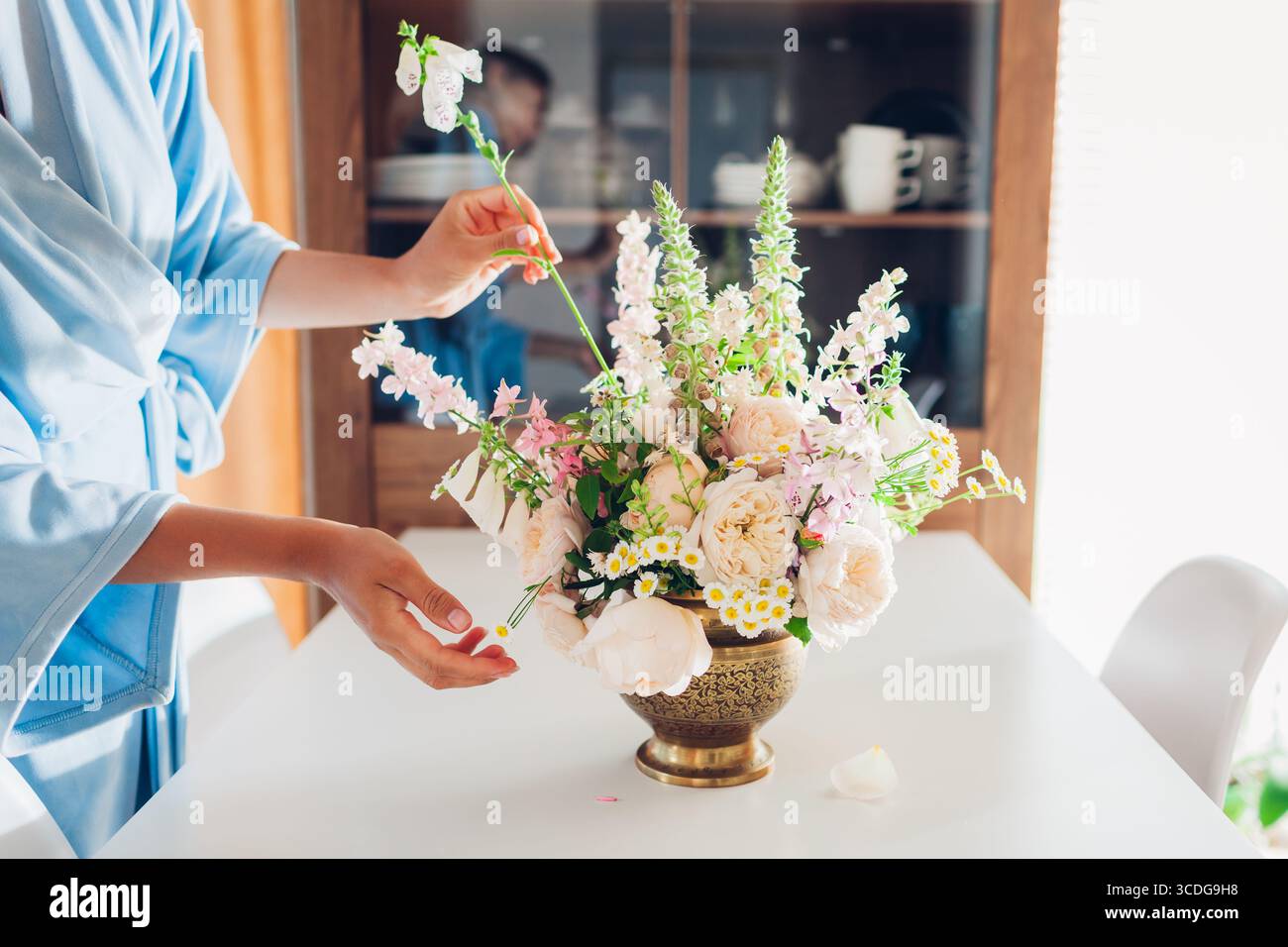 Frau, die Blumenstrauß in Messingvase voller Rosenblumen auf Esstisch für Schrank macht. Inneneinrichtung und Wohnkultur. Blumenarrangements im Sommer Stockfoto