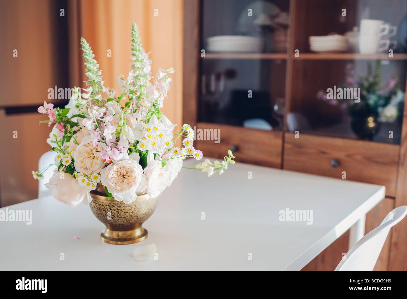 Frisches Blumenarrangement aus Messingvase auf Esstisch zur Dekoration der Kücheneinrichtung. Blumenstrauß aus weißen, beigen Rosen, Digitalis blüht links Stockfoto