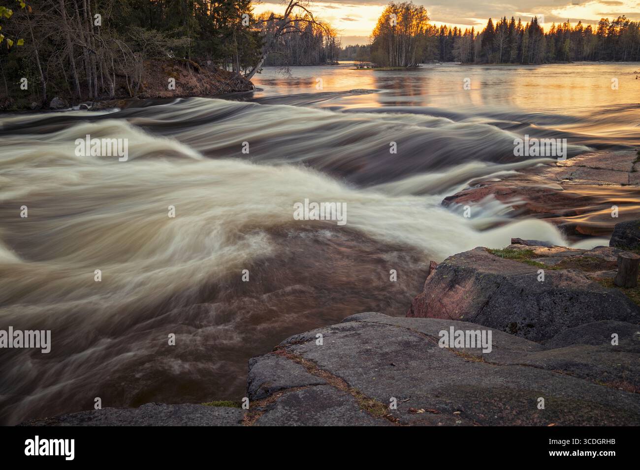 Eine Langzeitaufnahme des Kymijoki-Flusses, Pernoonkosket, zeigt die seidige Bewegung des Wassers, das bei Sonnenuntergang über Felsen rauscht. Der Himmel ist ein warmer golde Stockfoto