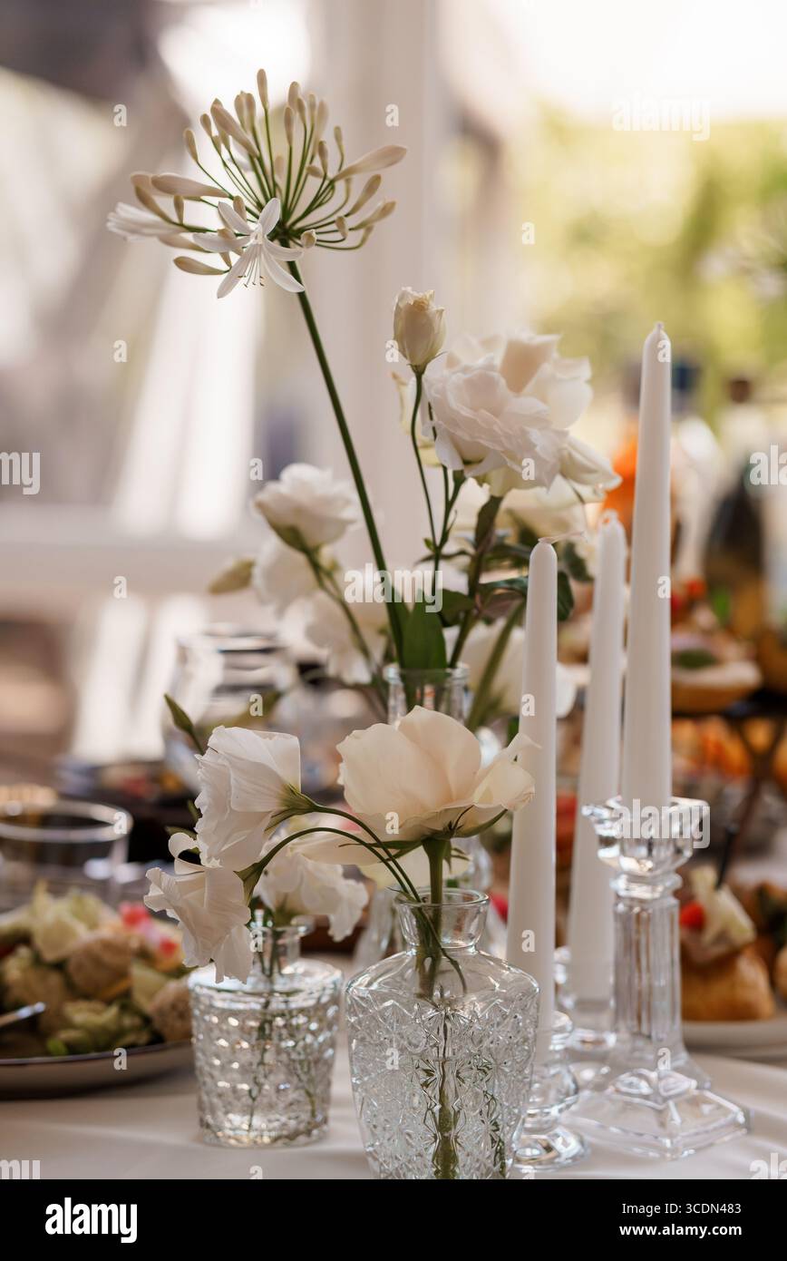 Ein wunderschön gestalteter Hochzeitstisch bietet zarte weiße Rosen, süße Erbsen und markante Spinnenlilien in Vintage-Kristallvasen neben schlanken Weißweinen Stockfoto