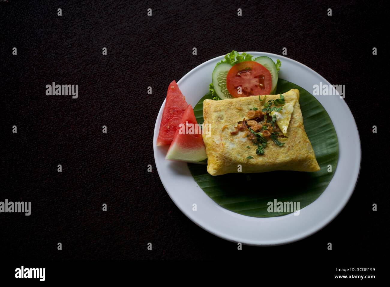 Köstlicher gebratener Reis in einem quadratischen Omelette auf Bananenblättern, garniert mit Sellerieblättern, gebratenen Schalotten, Gurkenscheiben, in Scheiben geschnittenen Wassermelonen A Stockfoto