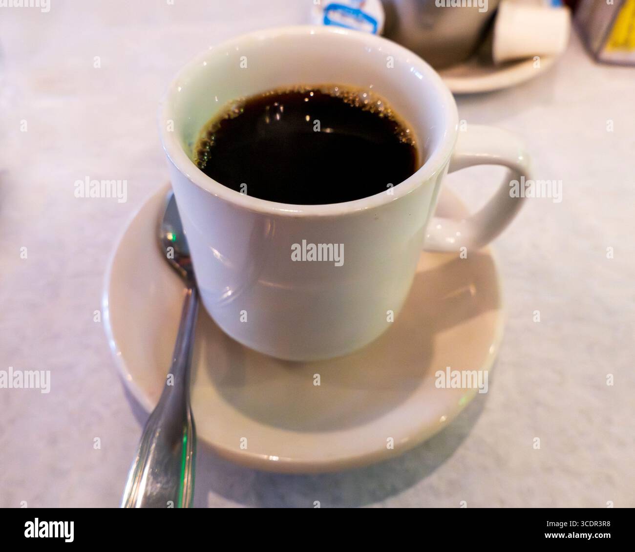 Eine dampfende Tasse schwarzen Kaffees steht auf einer weißen Untertasse mit einem Löffel, bereit zum Genießen. Die tiefe, reichhaltige Flüssigkeit hat einen leichten Schaum auf der Oberseite, der zu einem Wa einlädt Stockfoto