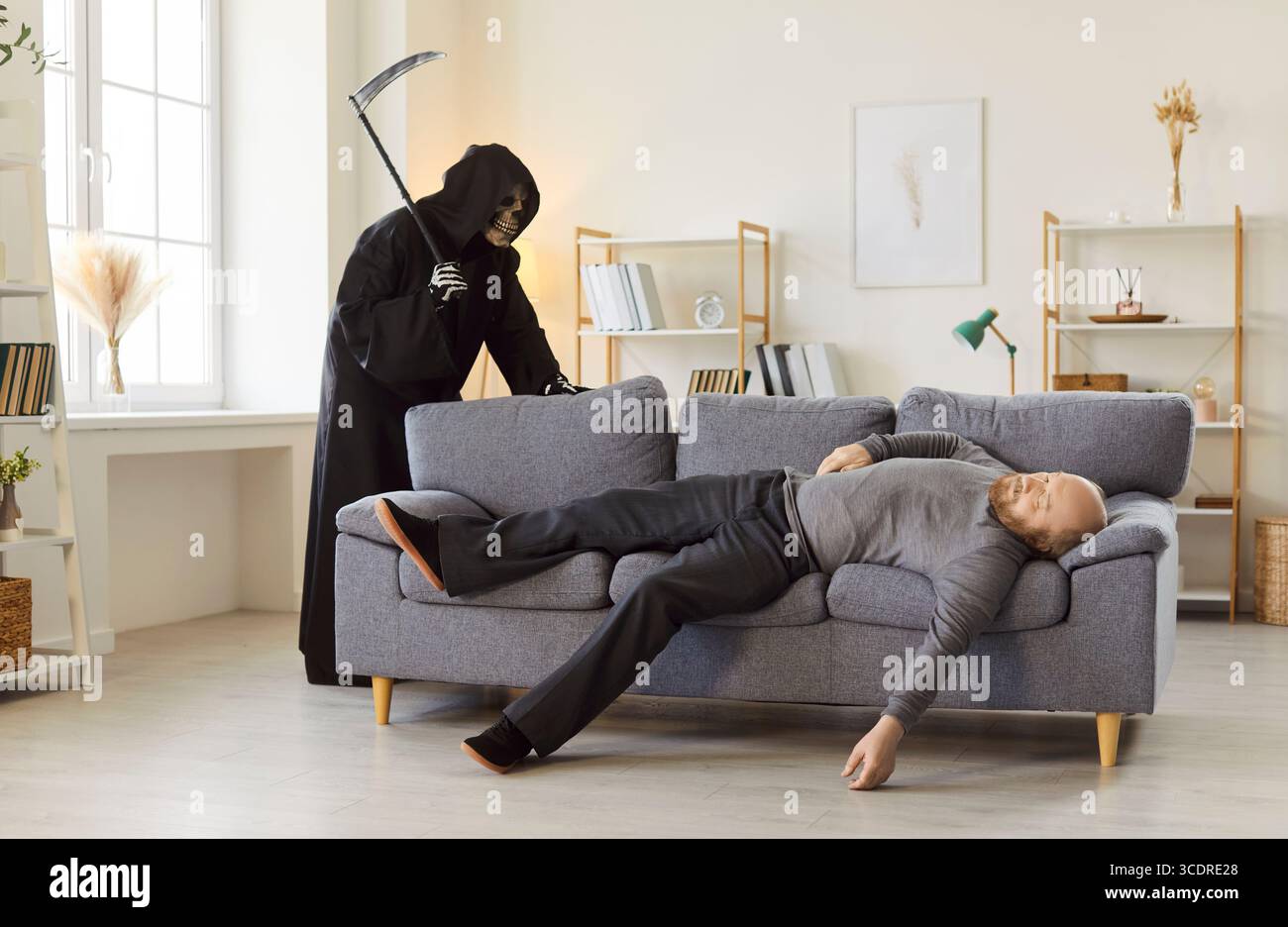 Finstere Figur eines grimmigen Sensers des Todes mit Sense, die hinter leblosen Mann auf Couch steht. Stockfoto