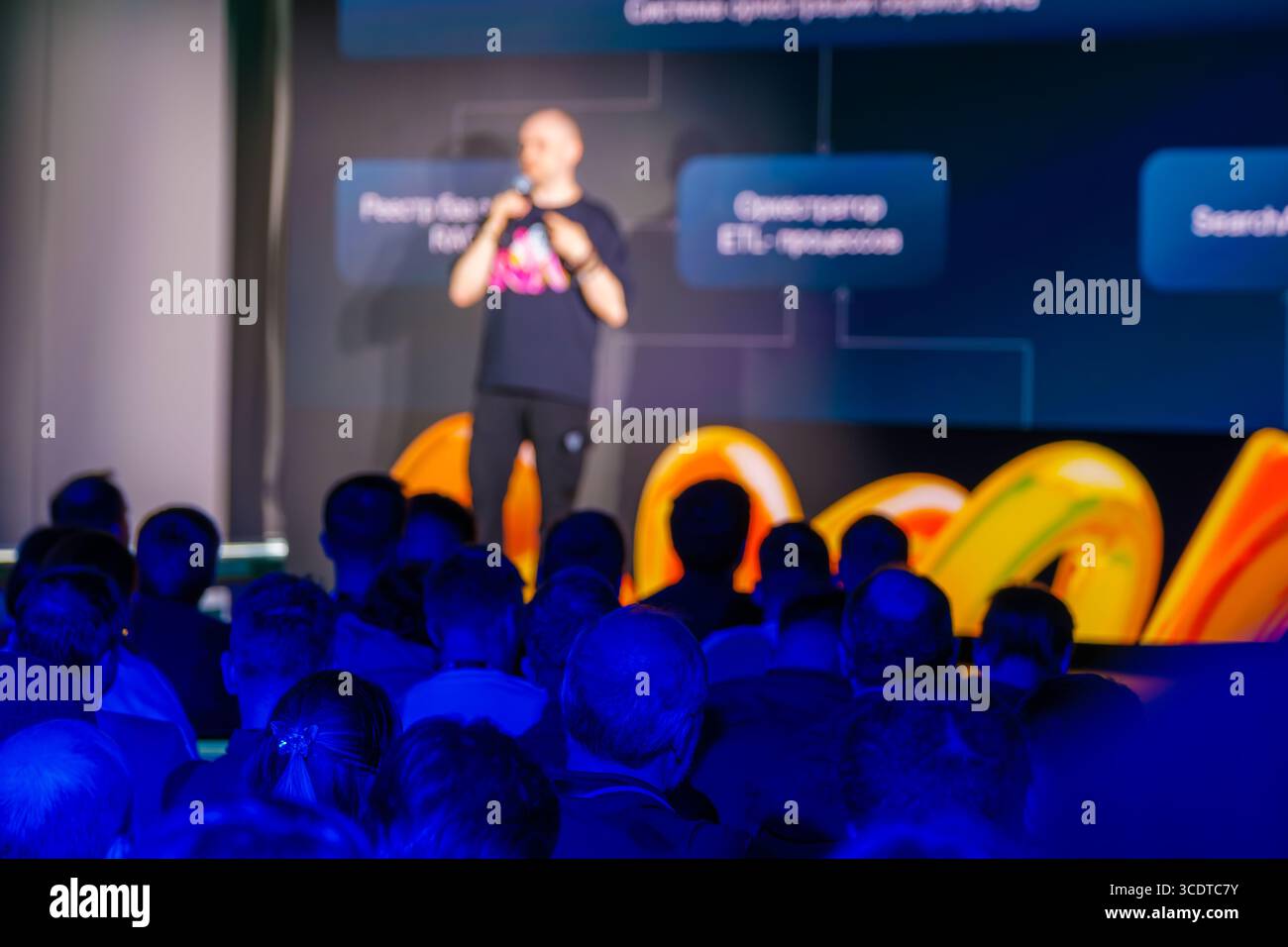 Professioneller Referent auf der Bühne einer Konferenz mit gezielter Beteiligung des Publikums. Der Raum ist mit lebhaften Farben beleuchtet und bietet eine dynamische Umgebung zum Austausch von Ideen und Networking. Stockfoto