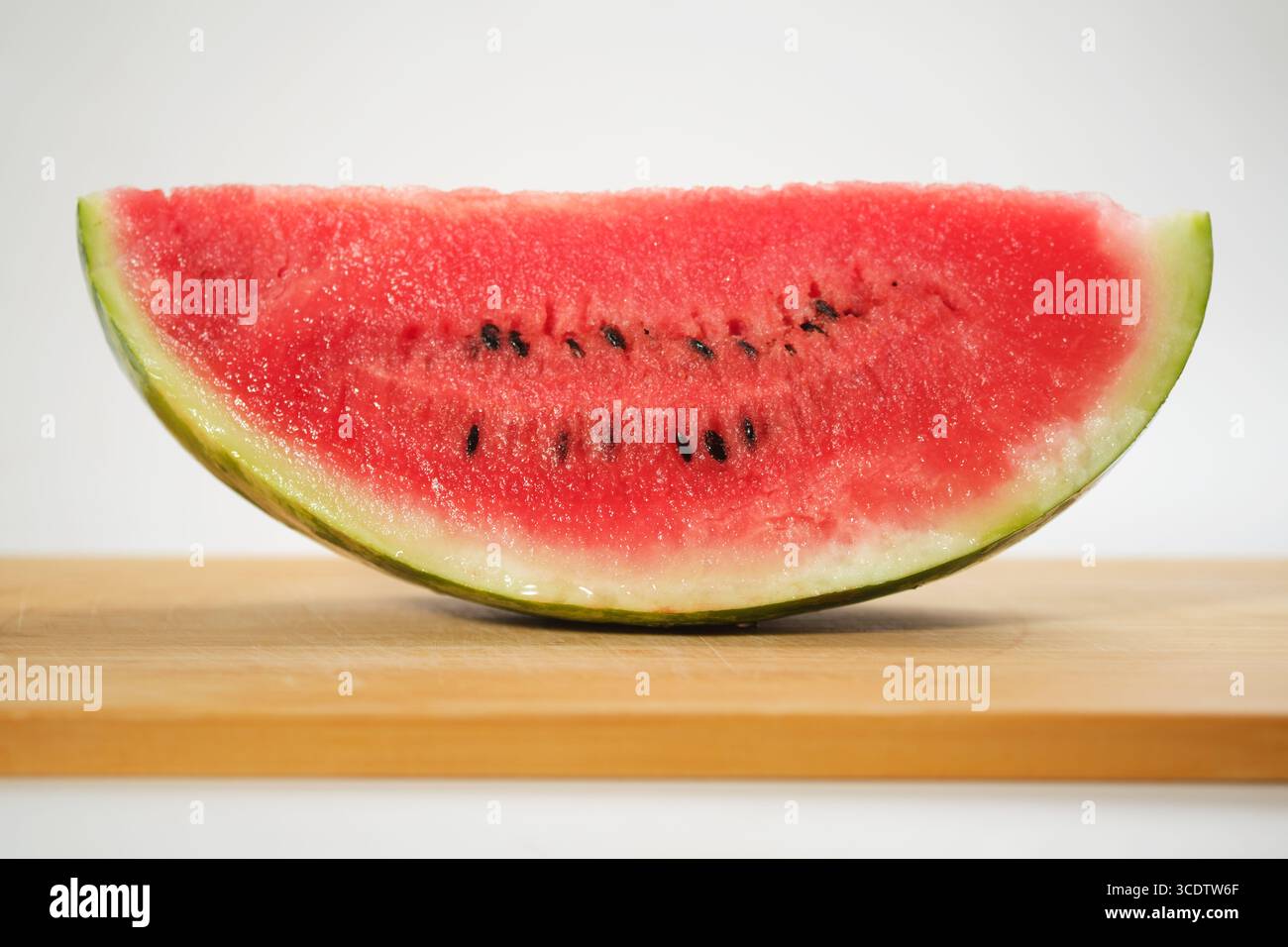 Ein Nahaufnahme einer Wassermelonenscheibe auf einem Holzbrett vor weißem Hintergrund, die seine saftige Textur und leuchtende rote Farbe zeigt Stockfoto