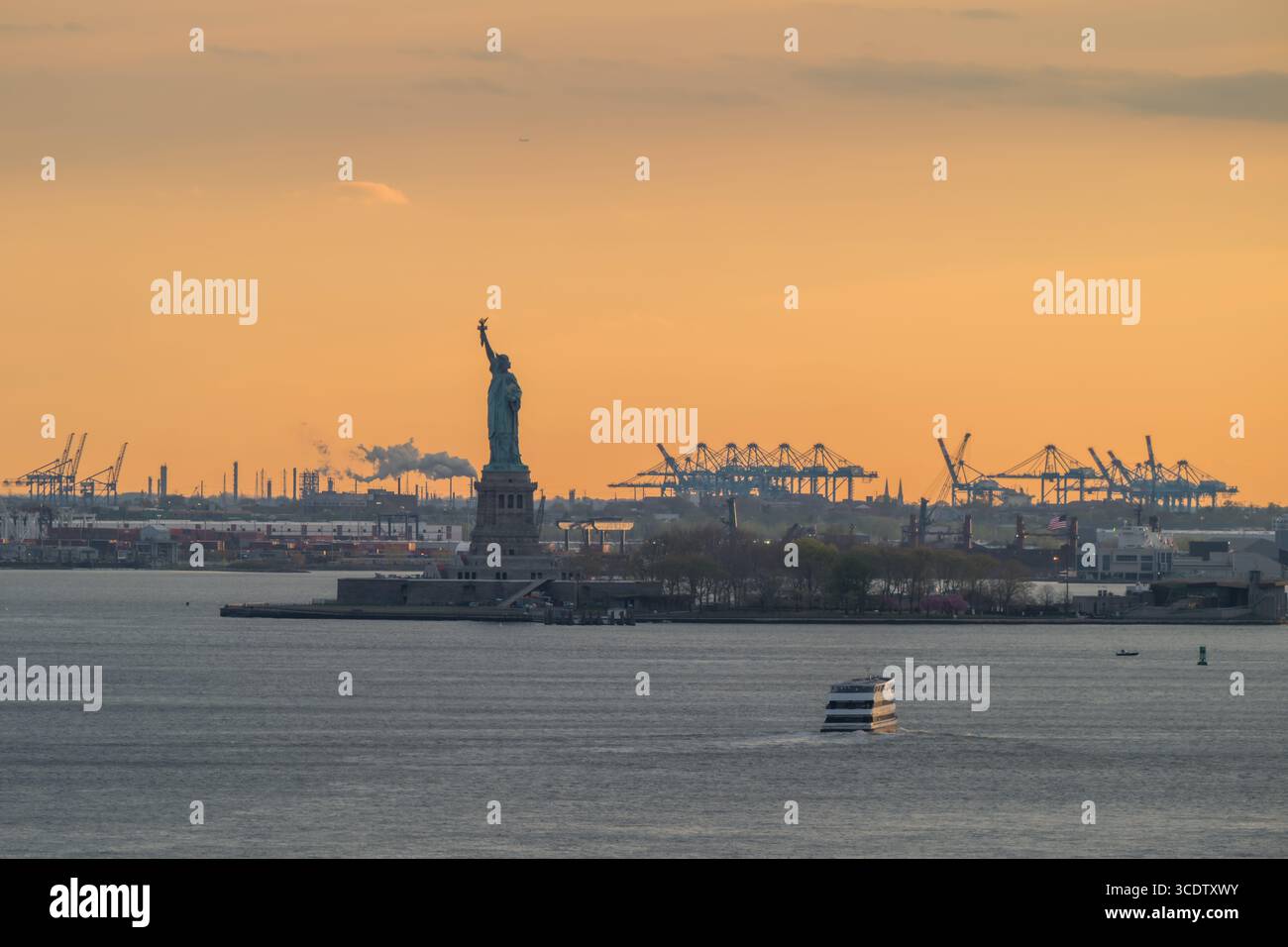 Blick auf Lady Liberty steht hoch vor dem feurigen Kuss des Sonnenuntergangs, ihre Silhouette ein Leuchtfeuer der Freiheit inmitten des geschäftigen Hafens, New York, New York Stockfoto
