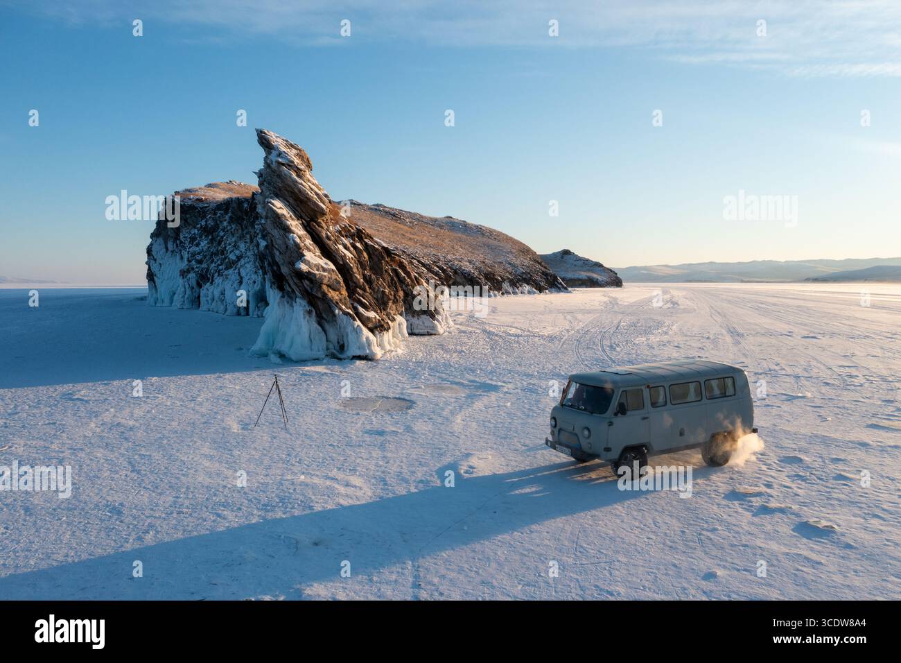 Der Blick aus der Vogelperspektive auf einen zerklüfteten Felsvorsprung mit eisigen Formationen steht im Kontrast zu der riesigen, schneebedeckten Weite, wo ein Van die gefrorene Landschaft, den Baikalsee, in Russland, durchquert. Stockfoto
