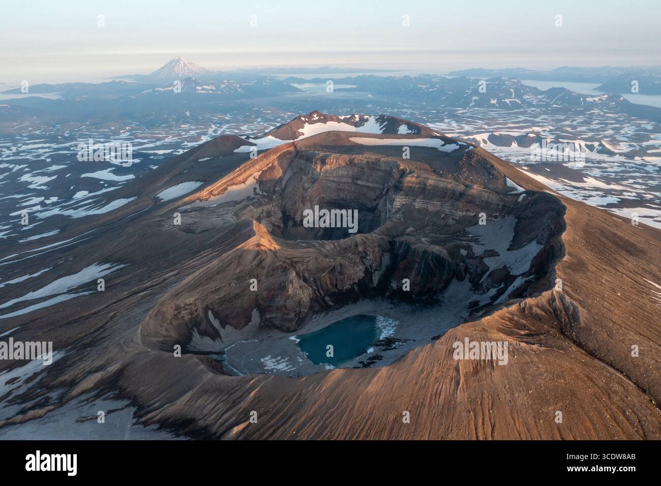 Der Blick aus der Vogelperspektive auf die zerklüftete Caldera des Gorely Volcano mit einem türkisfarbenen See im Inneren steht in starkem Kontrast zu den weit entfernten, schneebedeckten Gipfeln und Stockfoto