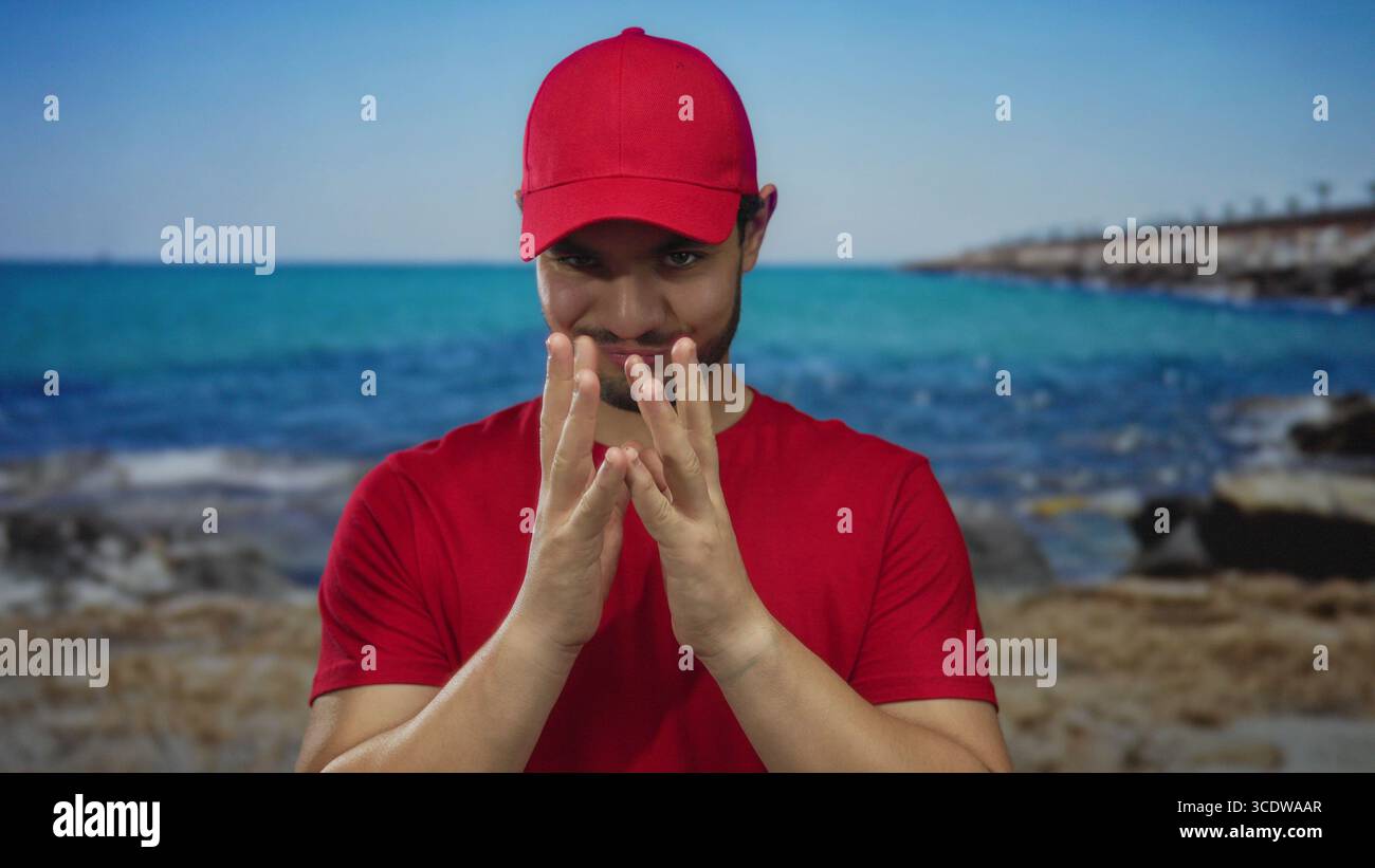 Junger hispanischer Kuriermann in roter Mütze und Hemd, mit den Fingern geschwenkt und mit einem leisen Grinsen an einem Strandpier. Stockfoto