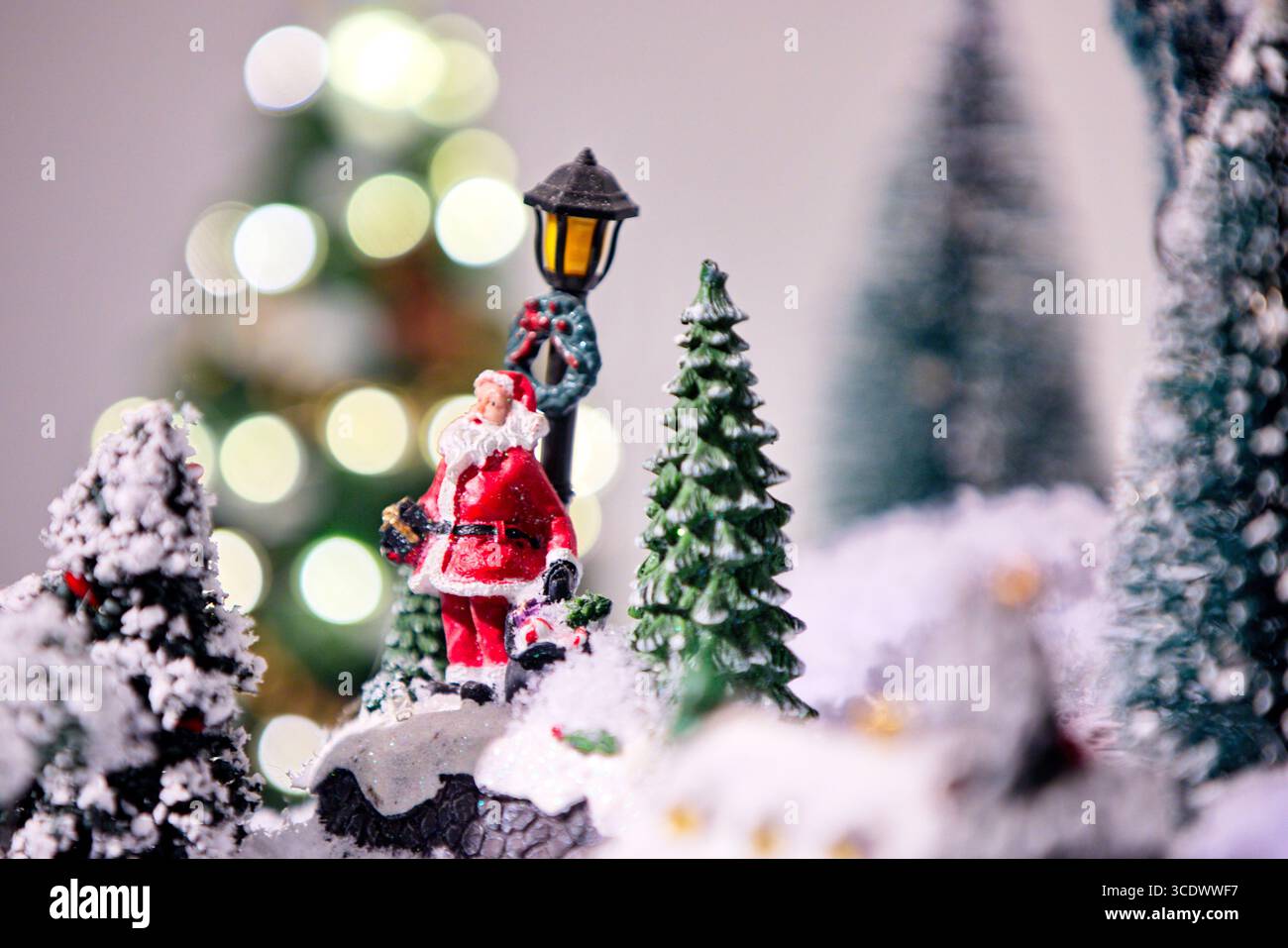 Miniatur-Weihnachtsmann-Figur mit Kranz an einem Laternenpfahl in einer verschneiten Dorfszene, festlichen Bokeh-Lichtern und Bäumen dahinter, ein Studio-Stillleben für die Lizenzierung. Stockfoto